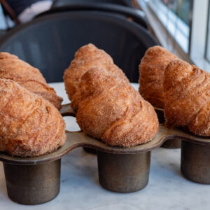 The morning bun is our house-made croissant dough that is rolled and baked in muffin pans. Once baked, we toss the morning buns in a mixture of cardamom, cinnamon, and sugars. Baked fresh daily.